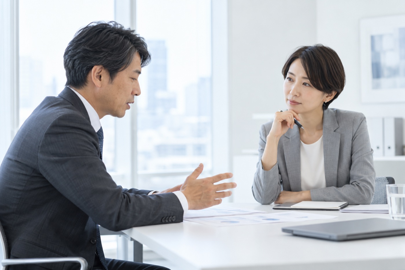 明るい会議室で、スーツ姿の男性が身ぶりを交えて話し、向かいの女性がペンを手に静かに耳を傾けている面談の様子。