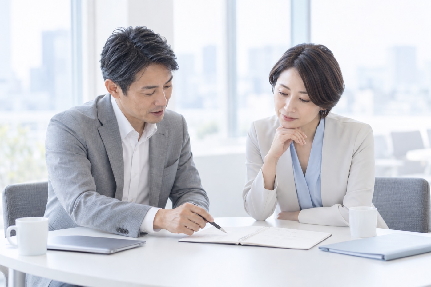明るいオフィスのテーブルで、男性がペンでノートを指しながら話し、隣の女性が頬に手を添えてそれを見ている。