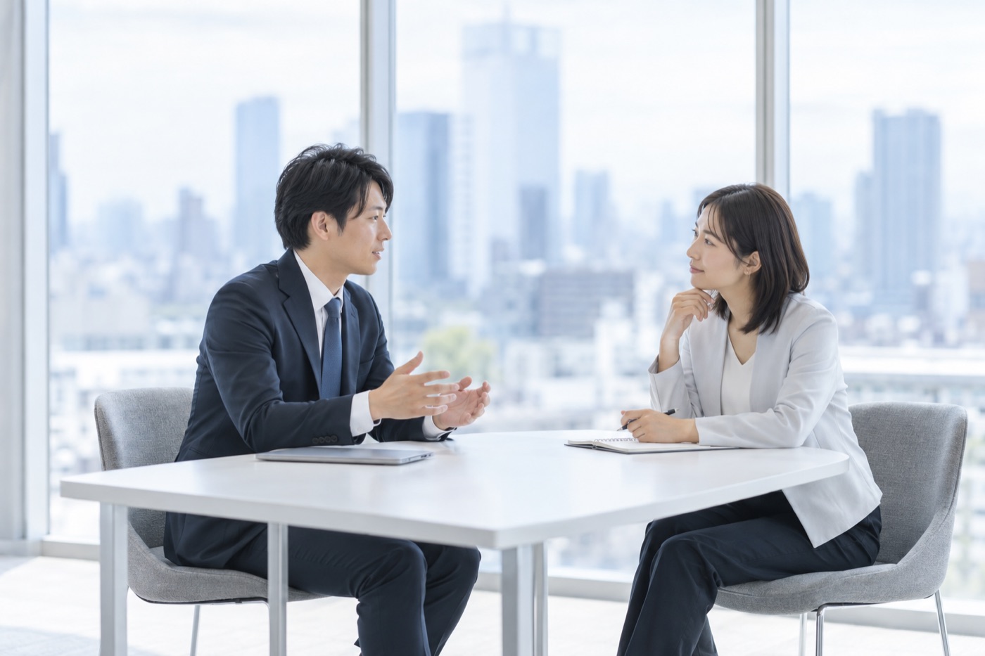 明るいオフィスの窓際で、スーツ姿の男性が身ぶりを交えて話し、向かいの女性がペンを手に静かに耳を傾けている。