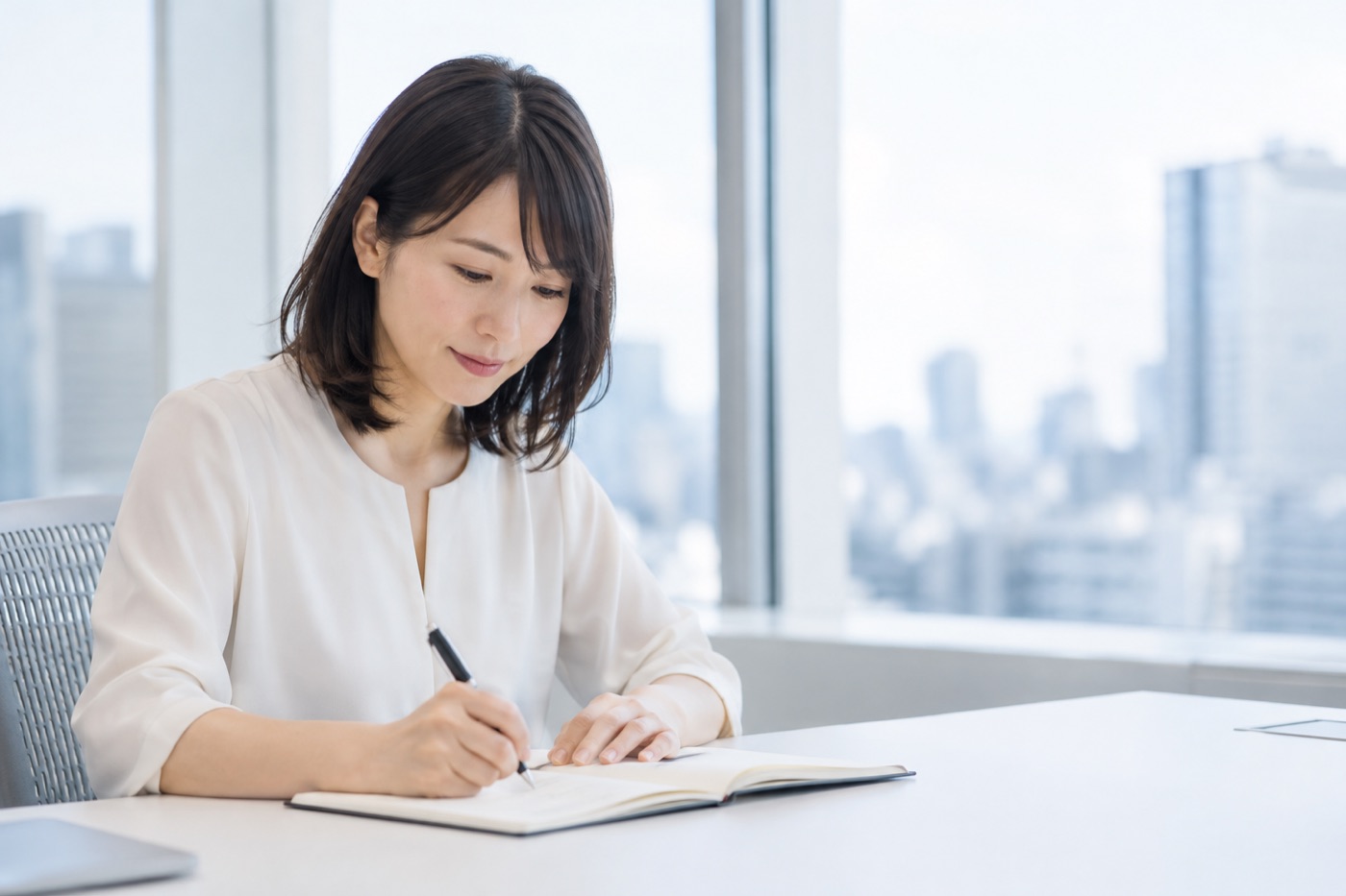 窓際の明るいデスクで、白いブラウスの女性が開いたノートにペンで書き込んでいる。窓の外にはビル街が広がっている。