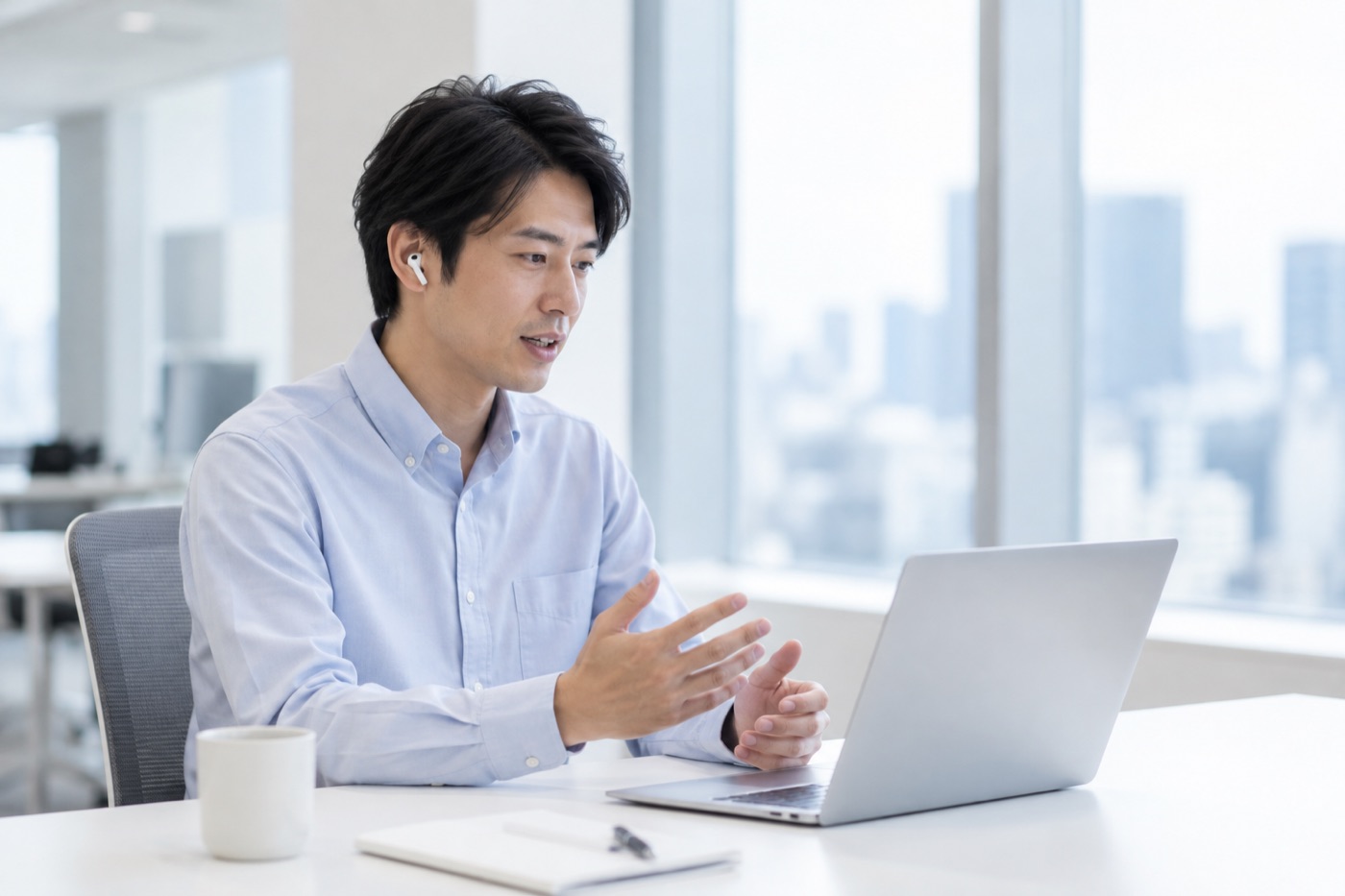 明るいオフィスで、イヤホンをつけた男性がノートパソコンに向かい、手を動かしながらオンラインで話している。